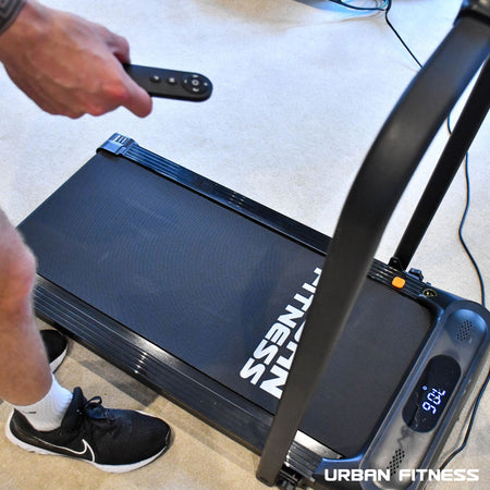 A person holding a remote control next to the Urban Fitness Walking Treadmill by Urban Fitness, ready for convenient workouts with smart fitness tracking.