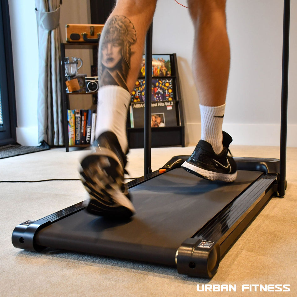 A person with a detailed calf tattoo walks indoors on the Urban Fitness Walking Treadmill, wearing white socks and black sneakers. Books and a camera are on shelves in the background. Urban Fitness smart tracking features are highlighted.