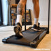 A person with a detailed calf tattoo walks indoors on the Urban Fitness Walking Treadmill, wearing white socks and black sneakers. Books and a camera are on shelves in the background. Urban Fitness smart tracking features are highlighted.