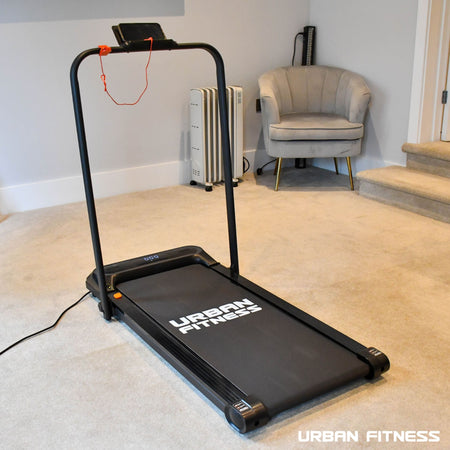 The Urban Fitness Walking Treadmill by Urban Fitness, in sleek black, is shown on a beige carpet at home near a gray armchair and heater, with a phone placed on its stand for smart fitness tracking.