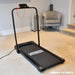 The Urban Fitness Walking Treadmill by Urban Fitness, in sleek black, is shown on a beige carpet at home near a gray armchair and heater, with a phone placed on its stand for smart fitness tracking.
