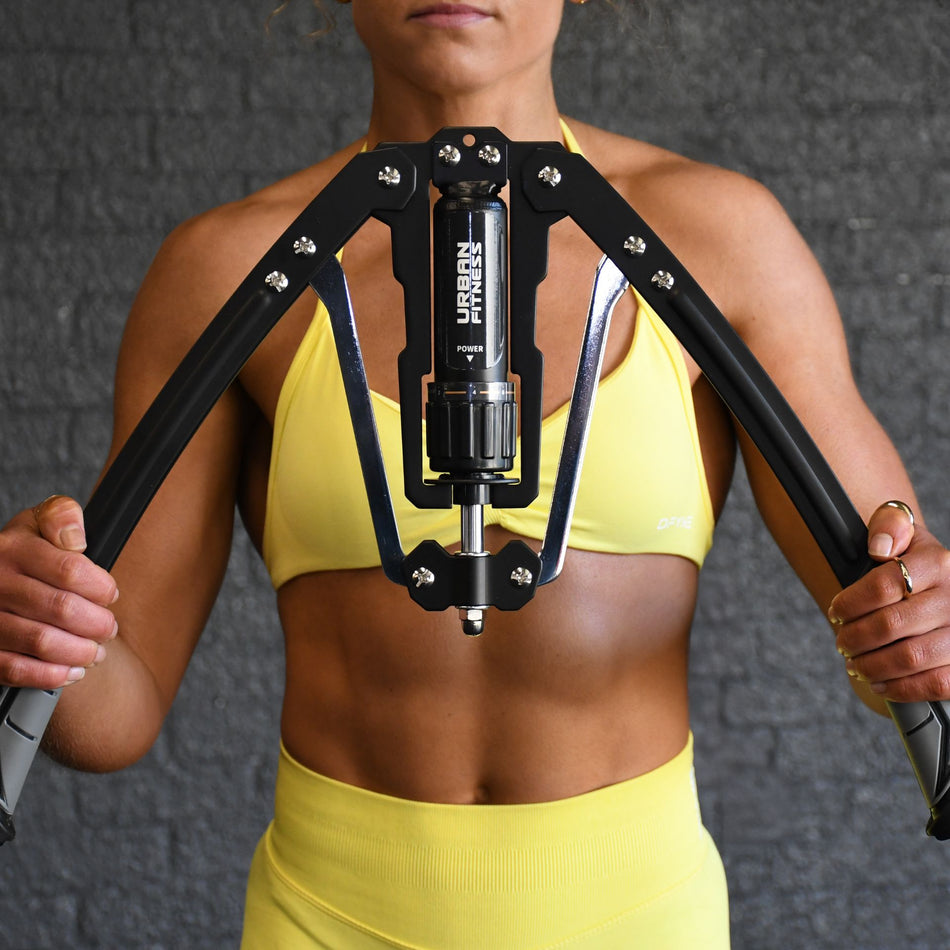 A woman in yellow sportswear uses the Urban Fitness Hydraulic Arms Trainer in Black/Grey, an adjustable resistance tool for upper body strength, with Urban Fitness branding, standing against a textured dark gray wall.