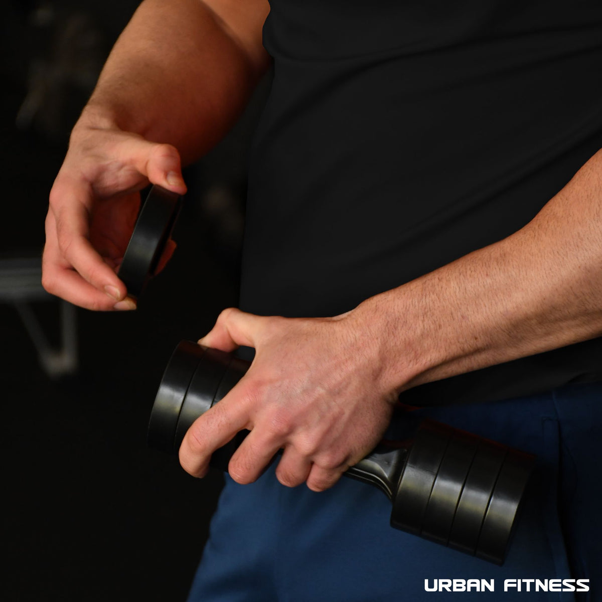 A person in a black shirt adjusts the quick-change weights on the Urban Fitness 2.5kg Adjustable Dumbbell Set in a gym, focusing on their hands and the equipment. The Urban Fitness logo appears at the bottom right corner of the image.