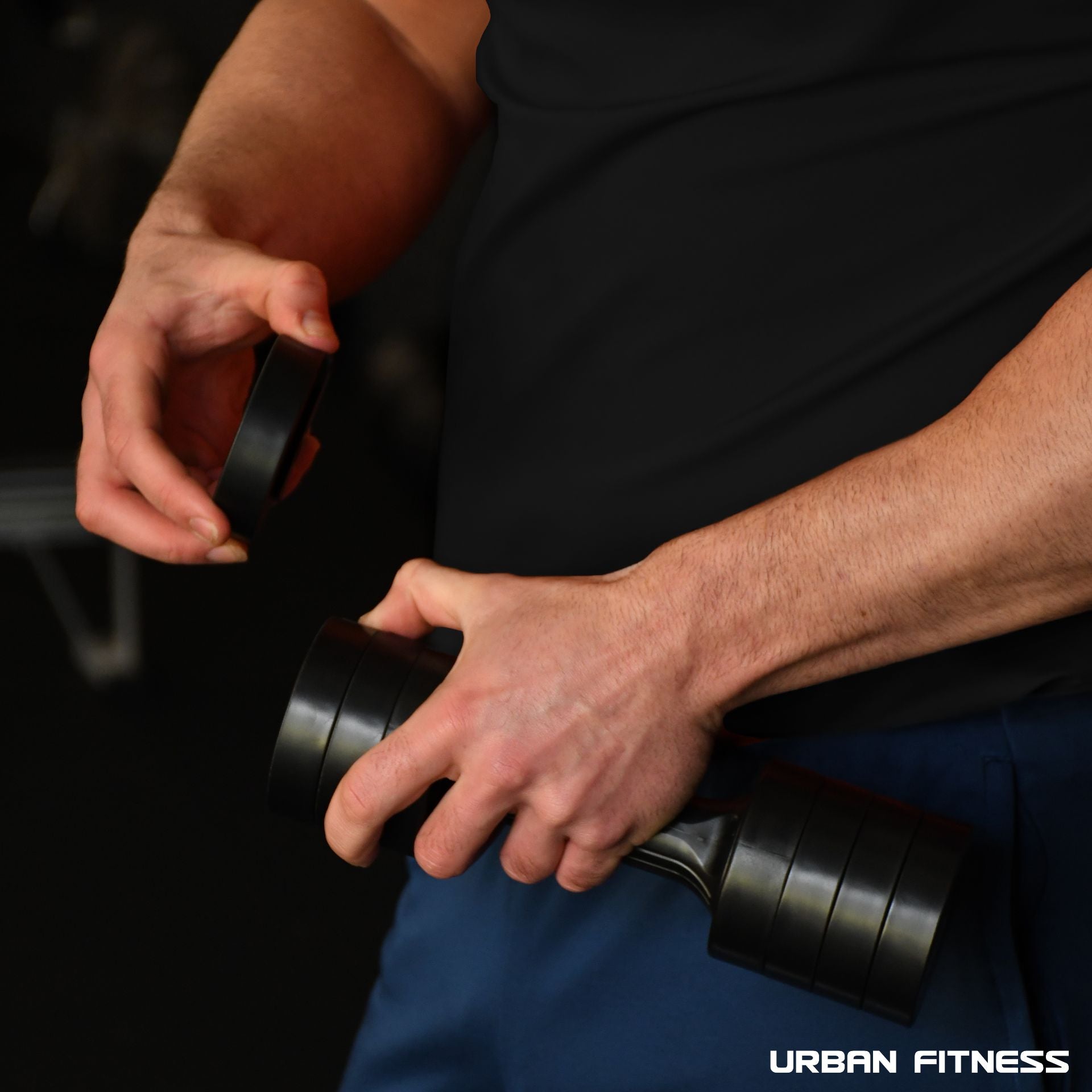 A person in a black shirt adjusts the quick-change weights on the Urban Fitness 2.5kg Adjustable Dumbbell Set in a gym, focusing on their hands and the equipment. The Urban Fitness logo appears at the bottom right corner of the image.