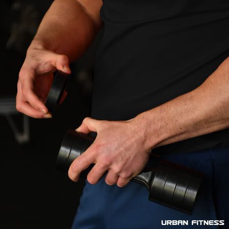 A person in a black shirt adjusts the quick-change weights on the Urban Fitness 2.5kg Adjustable Dumbbell Set in a gym, focusing on their hands and the equipment. The Urban Fitness logo appears at the bottom right corner of the image.