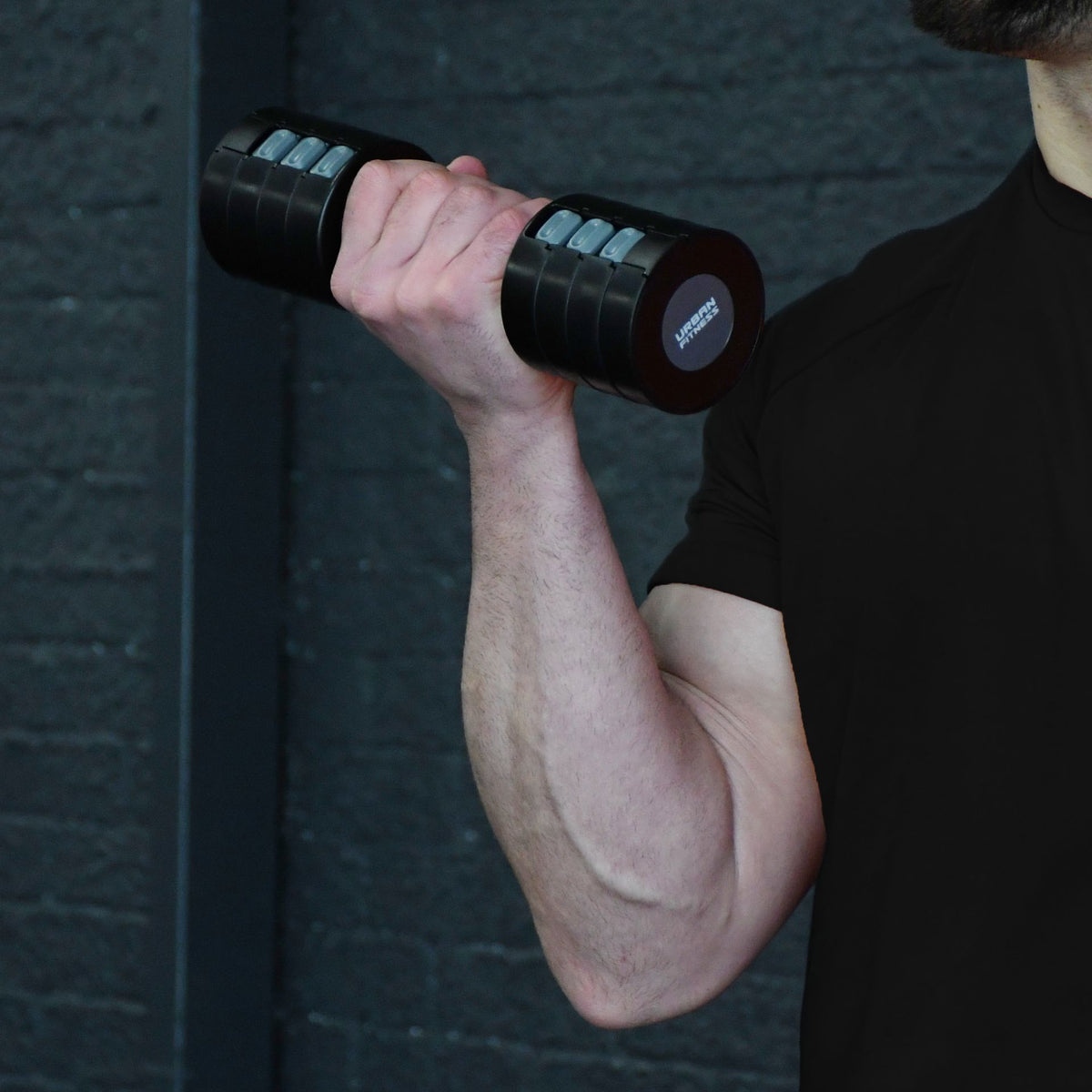 A person in a black shirt lifts the Urban Fitness 2.5kg Adjustable Dumbbell Set by Urban Fitness with their right arm against a dark, textured background.