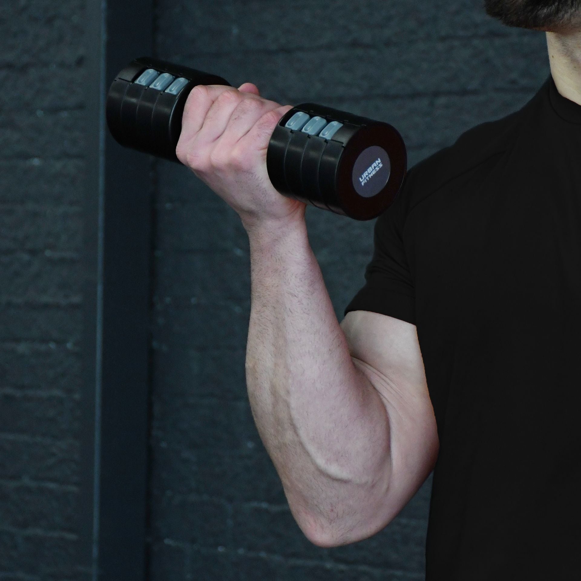 A person in a black shirt lifts the Urban Fitness 2.5kg Adjustable Dumbbell Set by Urban Fitness with their right arm against a dark, textured background.