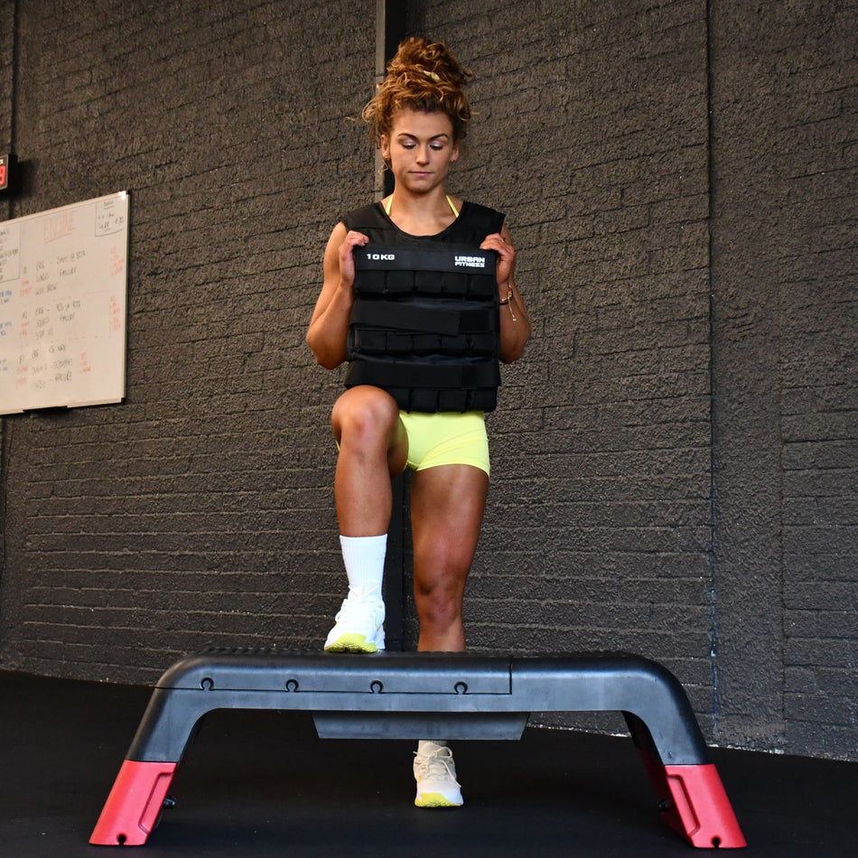 Wearing the Urban Fitness Adjustable Weighted Vest in black, a person steps onto an exercise platform in a gym for high-intensity conditioning, with one knee raised, a whiteboard, and gray walls visible in the background.