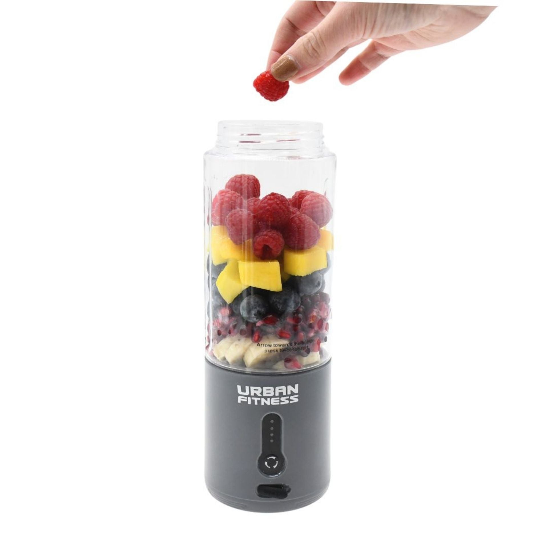 A hand adds a raspberry to the Urban Fitness VitalFuse Portable Blender, packed with raspberries, mango, blueberries, and banana. Rechargeable and printed with Urban Fitness branding, it's ideal for convenient on-the-go nutrition.
