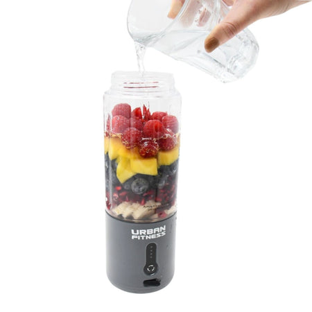 A hand pours water from a glass jug into the Urban Fitness VitalFuse Portable Blender filled with raspberries, mango, blueberries, bananas, and strawberries—an ideal on-the-go nutrition solution from Urban Fitness.