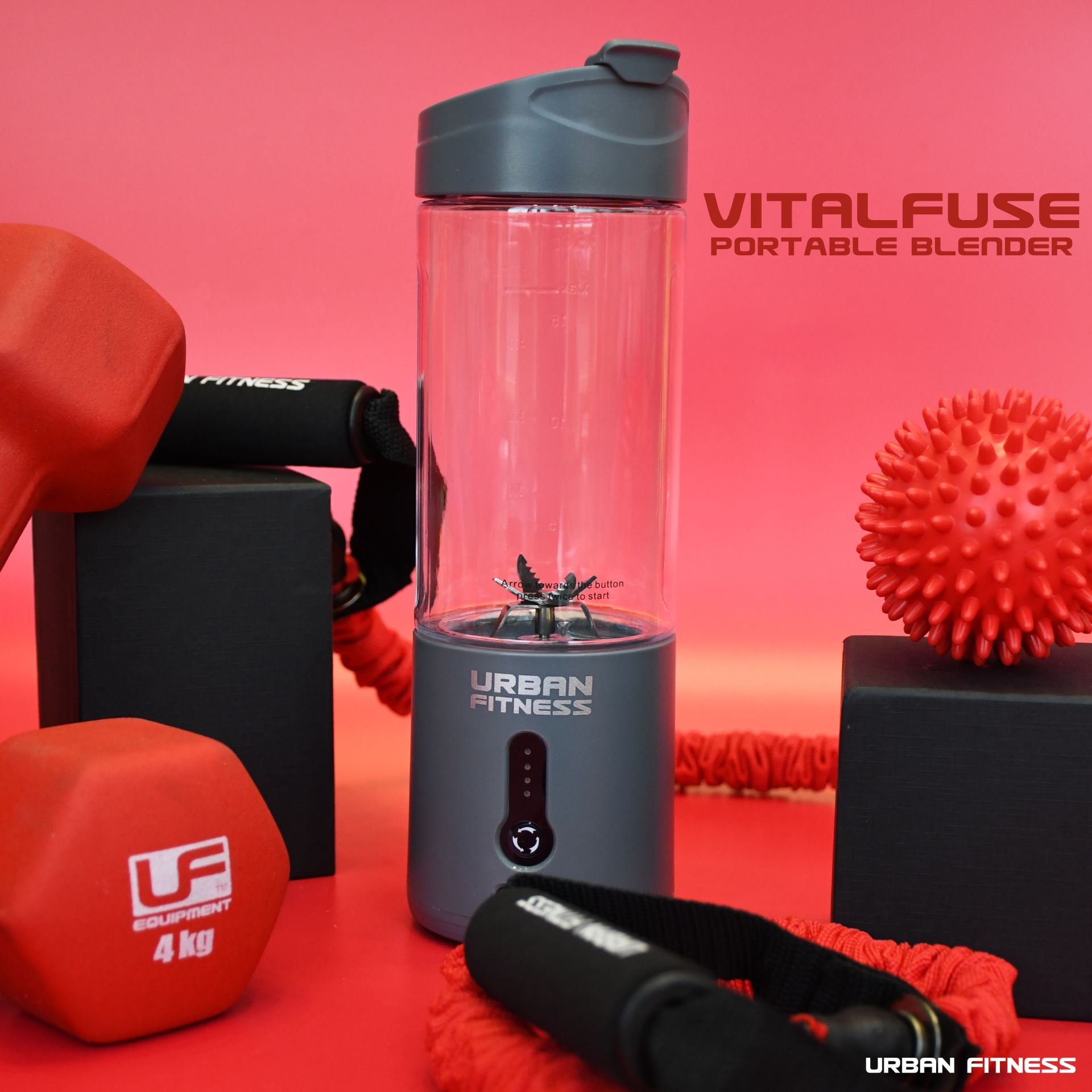 A grey Urban Fitness VitalFuse Portable Blender from Urban Fitness sits with red dumbbells, a spiky massage ball, resistance bands, and black fitness blocks on a red background—perfect for on-the-go nutrition. "VITALFUSE PORTABLE BLENDER" is in the top right.