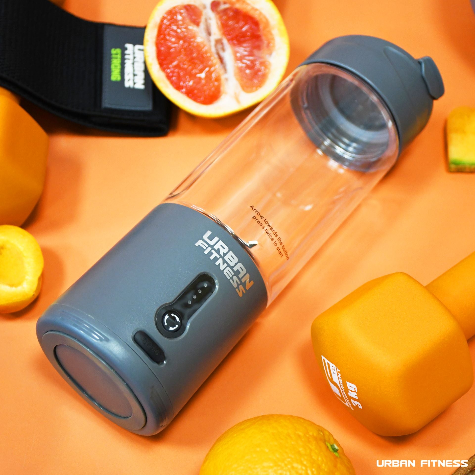 An Urban Fitness VitalFuse Portable Blender rests on an orange surface with orange dumbbells, a sliced grapefruit, and an orange nearby, highlighting vibrant on-the-go nutrition and a health-conscious lifestyle.