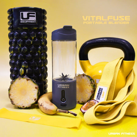 A black foam roller, yellow kettlebell, resistance band, half a pineapple, banana, fitness cards, and the Urban Fitness VitalFuse Portable Blender with fruit inside are arranged on yellow—ideal for on-the-go nutrition. Text: Urban Fitness.