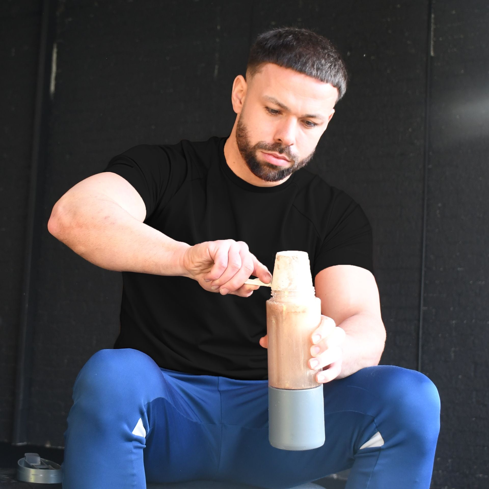 A man in a black shirt and blue pants scoops protein powder into the Urban Fitness VitalFuse Portable Blender by Urban Fitness, preparing convenient on-the-go nutrition against a dark background.