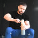 A man in a black shirt and blue pants scoops protein powder into the Urban Fitness VitalFuse Portable Blender by Urban Fitness, preparing convenient on-the-go nutrition against a dark background.