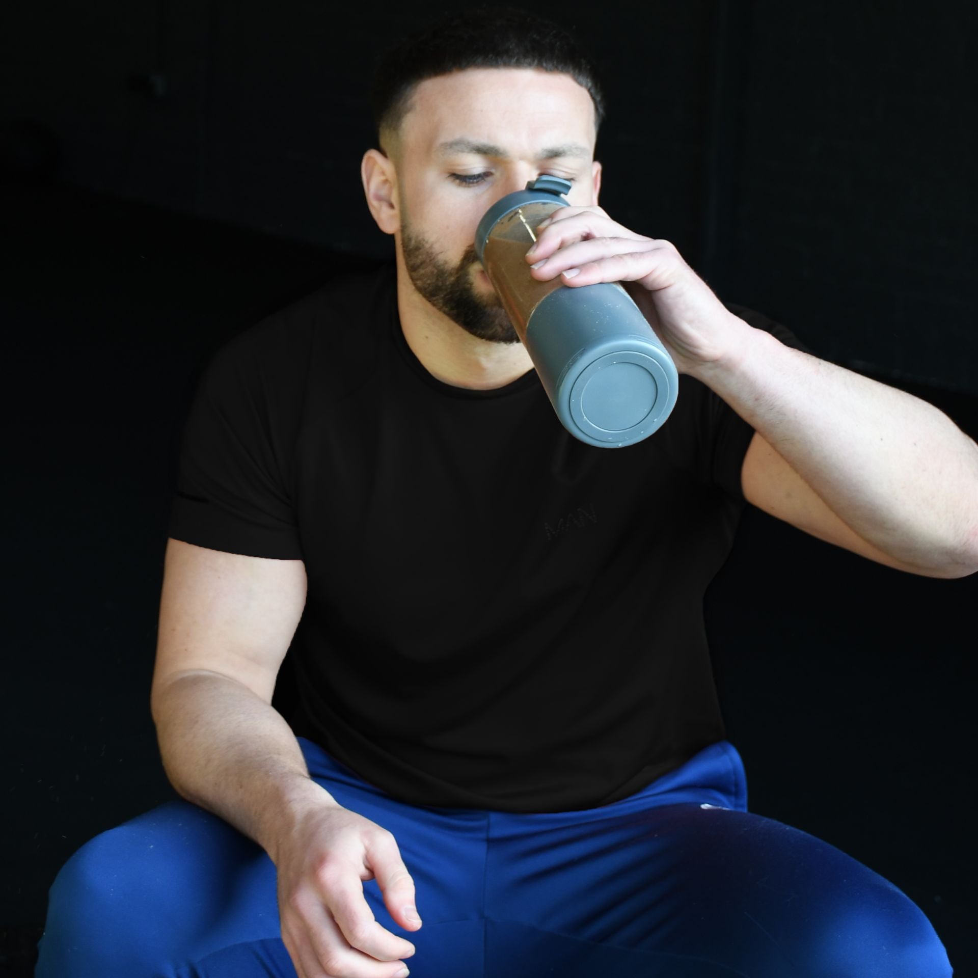 A man with short hair and a trimmed beard sits against a dark background, wearing a black t-shirt and blue pants, drinking from an Urban Fitness VitalFuse Portable Blender by Urban Fitness.