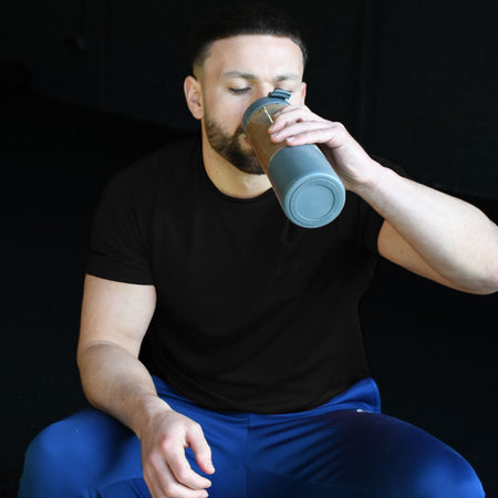A man with short hair and a trimmed beard sits against a dark background, wearing a black t-shirt and blue pants, drinking from an Urban Fitness VitalFuse Portable Blender by Urban Fitness.