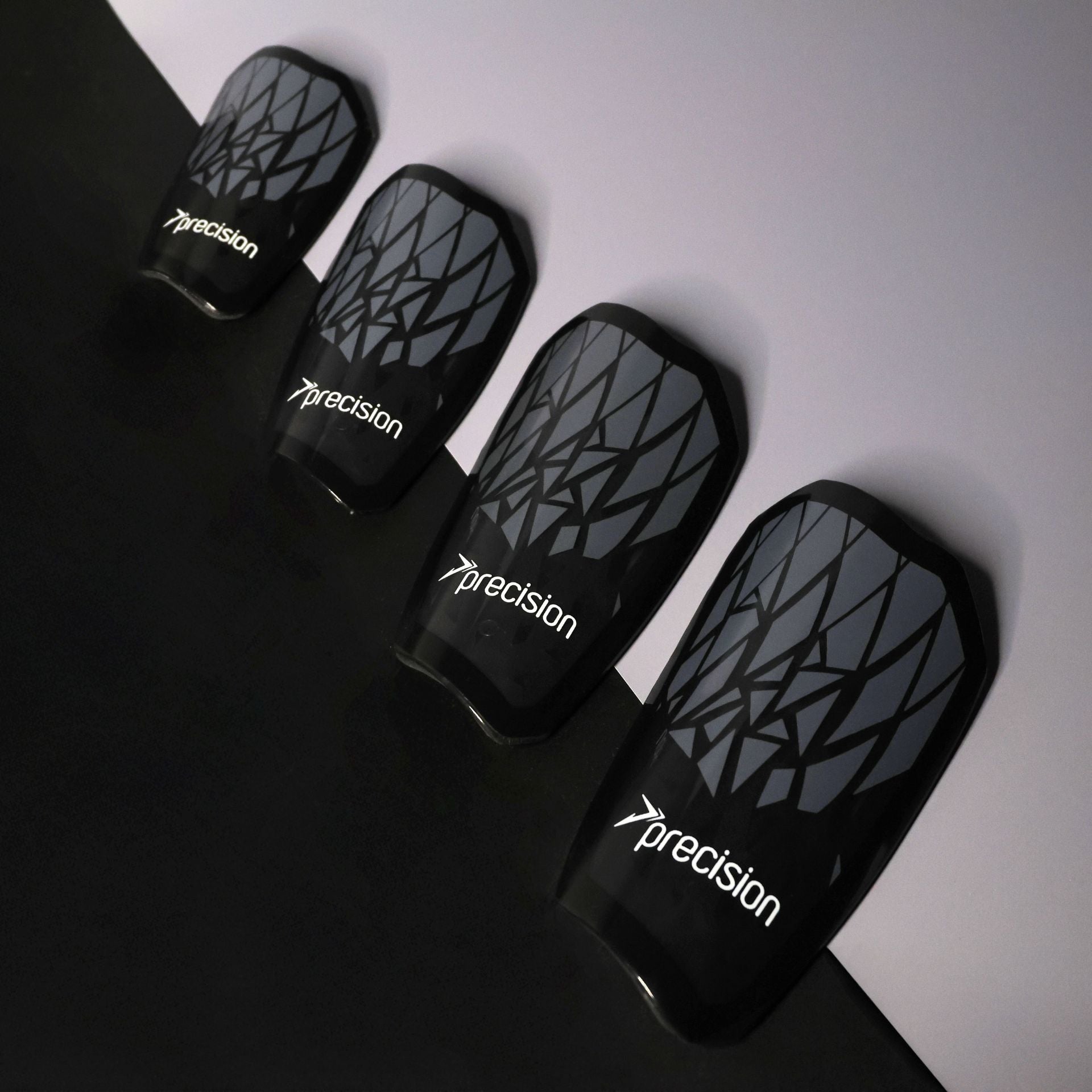 Four Precision Training Pro Fusion Flex Shin Guards in black with geometric patterns and "precision" printed on them are arranged diagonally on a two-tone black and gray background, delivering elite protection for athletes.