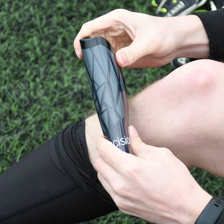 A person in black athletic gear adjusts Precision Training’s Precision Pro Fusion Flex Shin Guards, featuring a black and gray design, while seated on artificial turf to ensure elite protection for their next game.