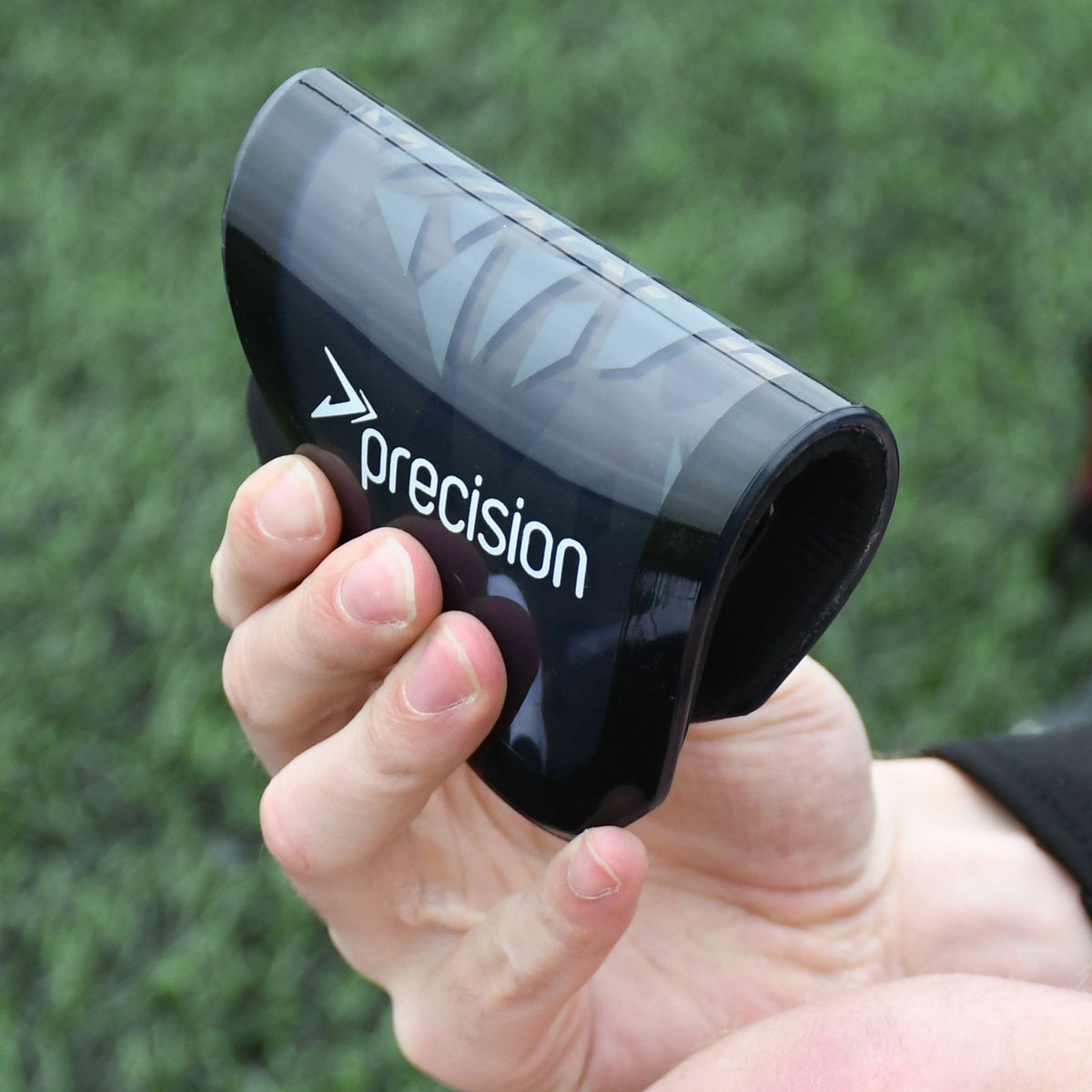 A person holds black Precision Pro Fusion Flex Shin Guards by Precision Training—these flexible guards offer elite protection, with artificial grass in the background.