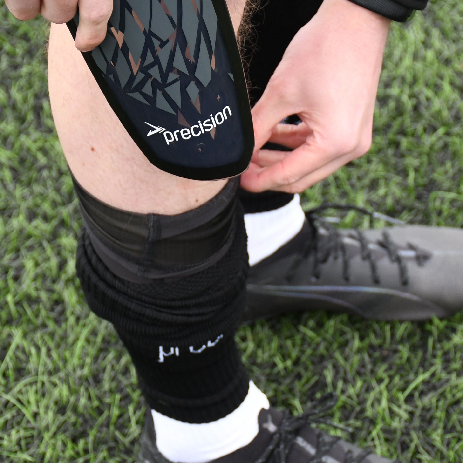 A person fits a Precision Training Pro Fusion Flex Shin Guard into their sock while wearing black football boots and standing on green artificial turf.
