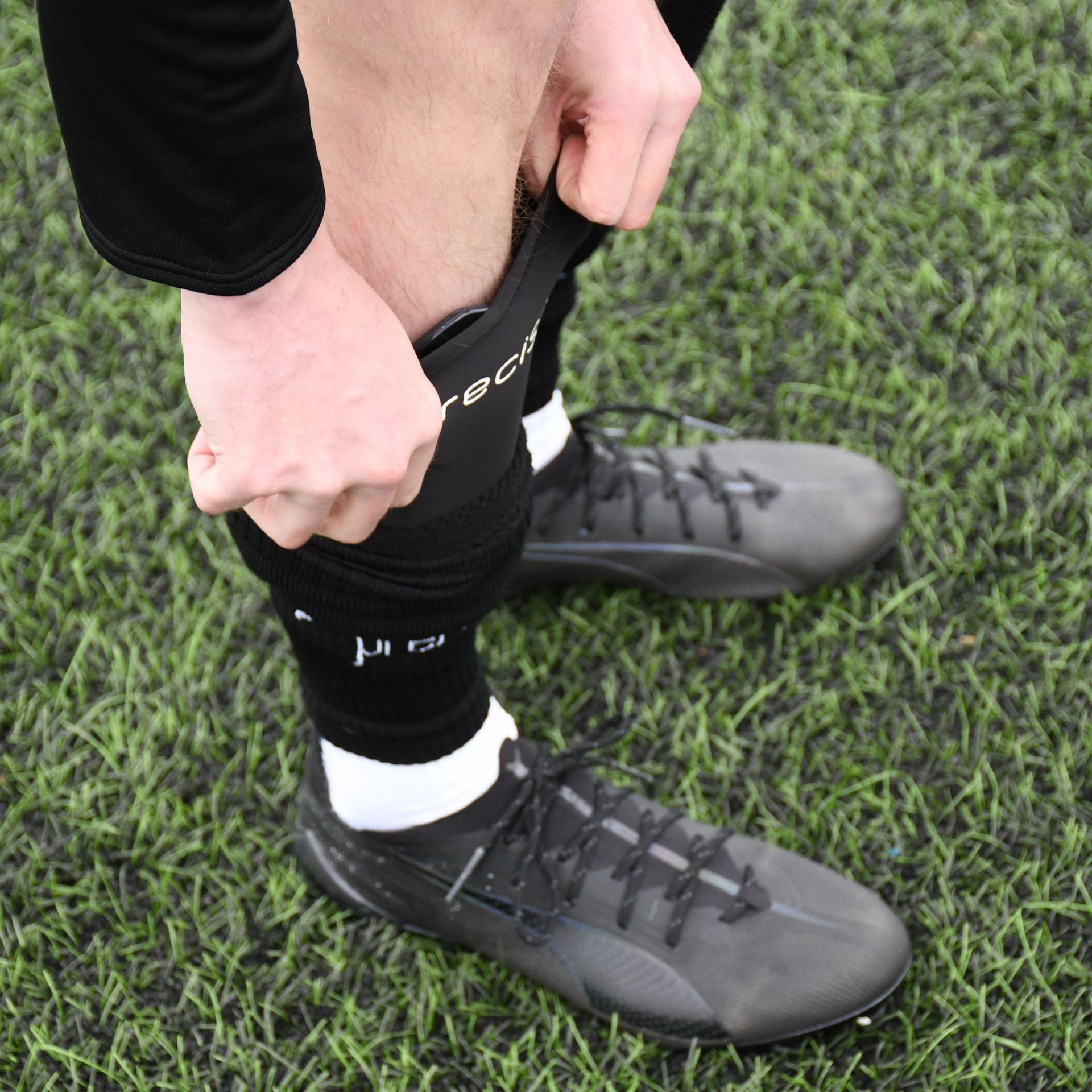 A player wearing black cleats and socks pulls up Precision Training’s Pro Fusion Flex Shin Guards while standing on green artificial turf.