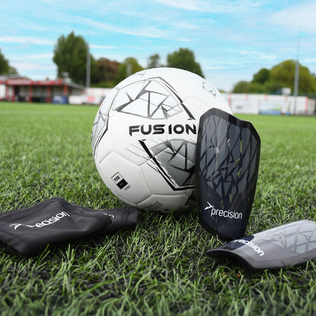 A Precision Pro Fusion Flex Shin Guard by Precision Training and a soccer ball sit on green artificial turf, with a stadium and trees in the background under a clear blue sky.