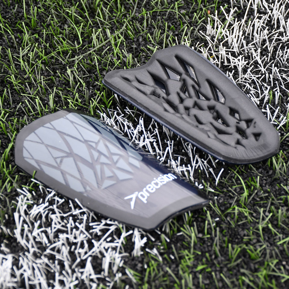 Two Precision Training Precision Pro Fusion Flex Shin Guards in black and grey with geometric patterns rest on artificial grass and white lines, one displaying the flexible exterior, the other showing its padded interior for elite protection.