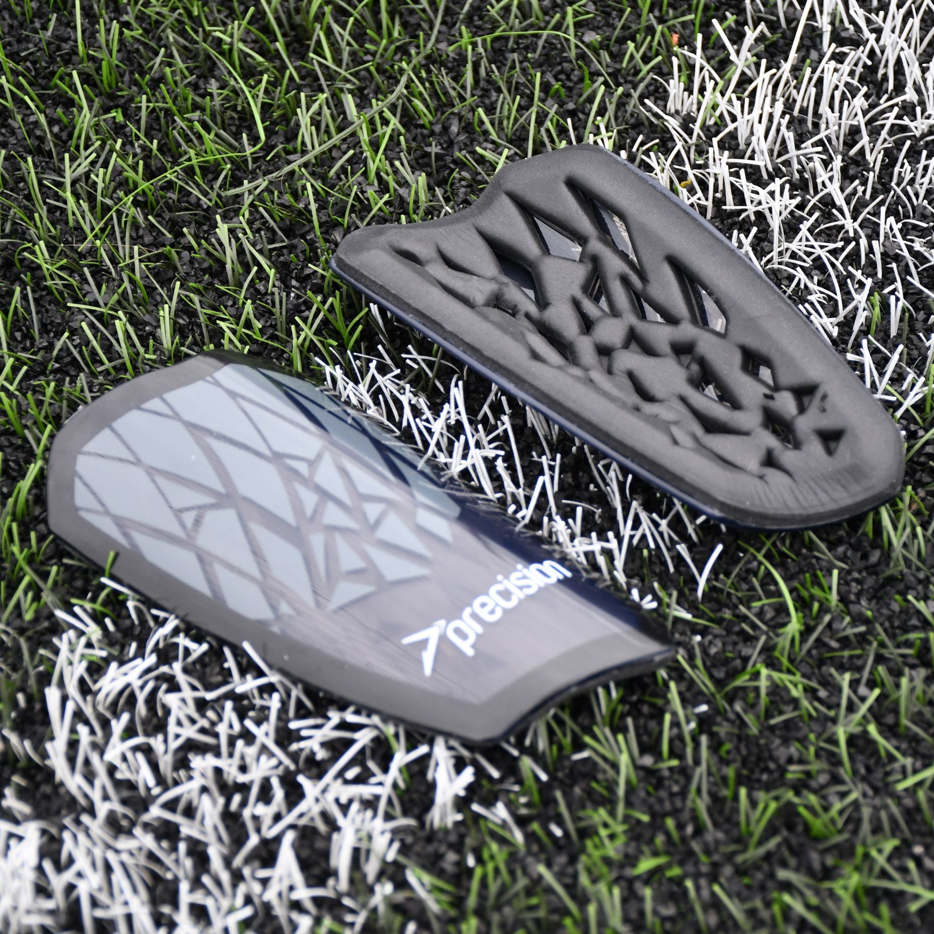 Two Precision Training Precision Pro Fusion Flex Shin Guards in black and grey with geometric patterns rest on artificial grass and white lines, one displaying the flexible exterior, the other showing its padded interior for elite protection.