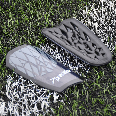 Two Precision Training Precision Pro Fusion Flex Shin Guards in black and grey with geometric patterns rest on artificial grass and white lines, one displaying the flexible exterior, the other showing its padded interior for elite protection.