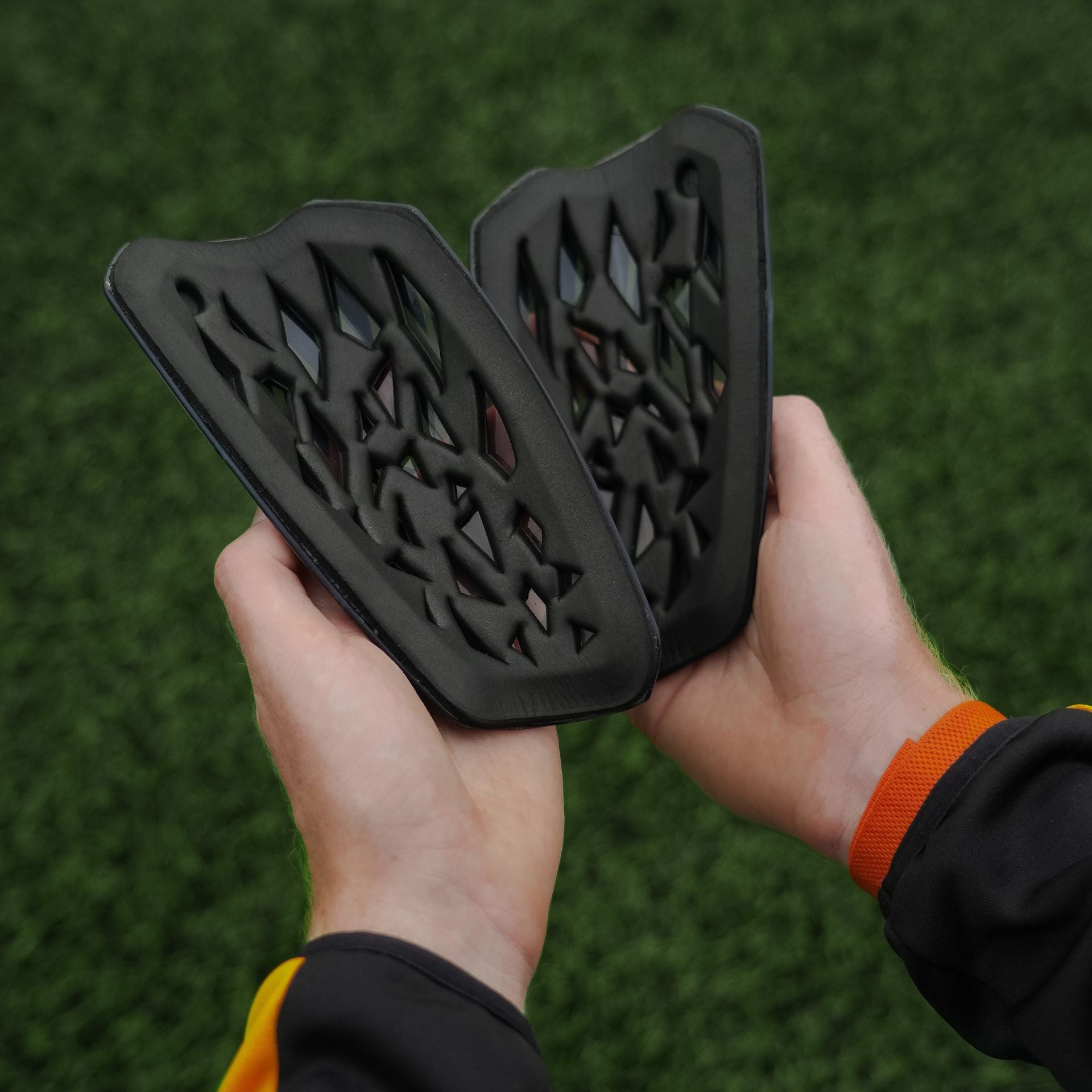A person holds two Precision Training Precision Pro Fusion Flex Shin Guards, black with a geometric cutout pattern, against blurred green artificial grass—designed for elite protection.
