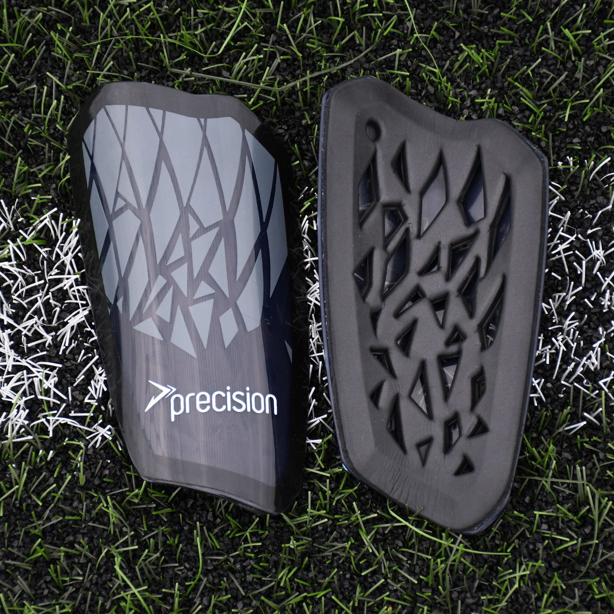 Two Precision Training Precision Pro Fusion Flex Shin Guards, black with a geometric pattern and "precision" on one, are placed side by side on artificial grass over a white field line, offering elite protection for soccer players.