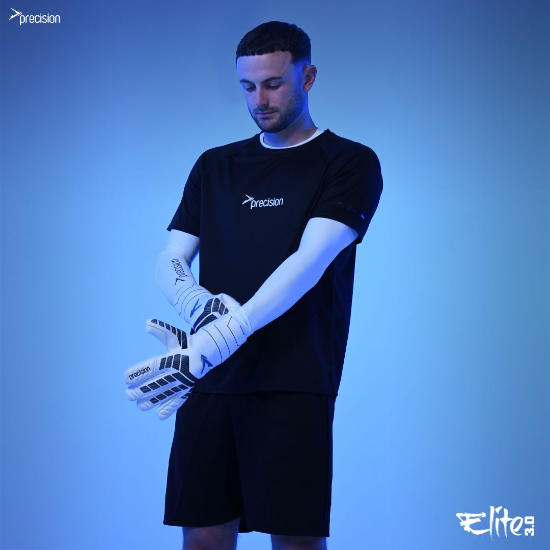 A man in a black t-shirt and shorts with Precision branding puts on Martin Berrill Sports Precision Elite 3.0 Contact Goalkeeper Gloves, renowned for their grip. The blue background displays Elite and Precision logos in the corners.