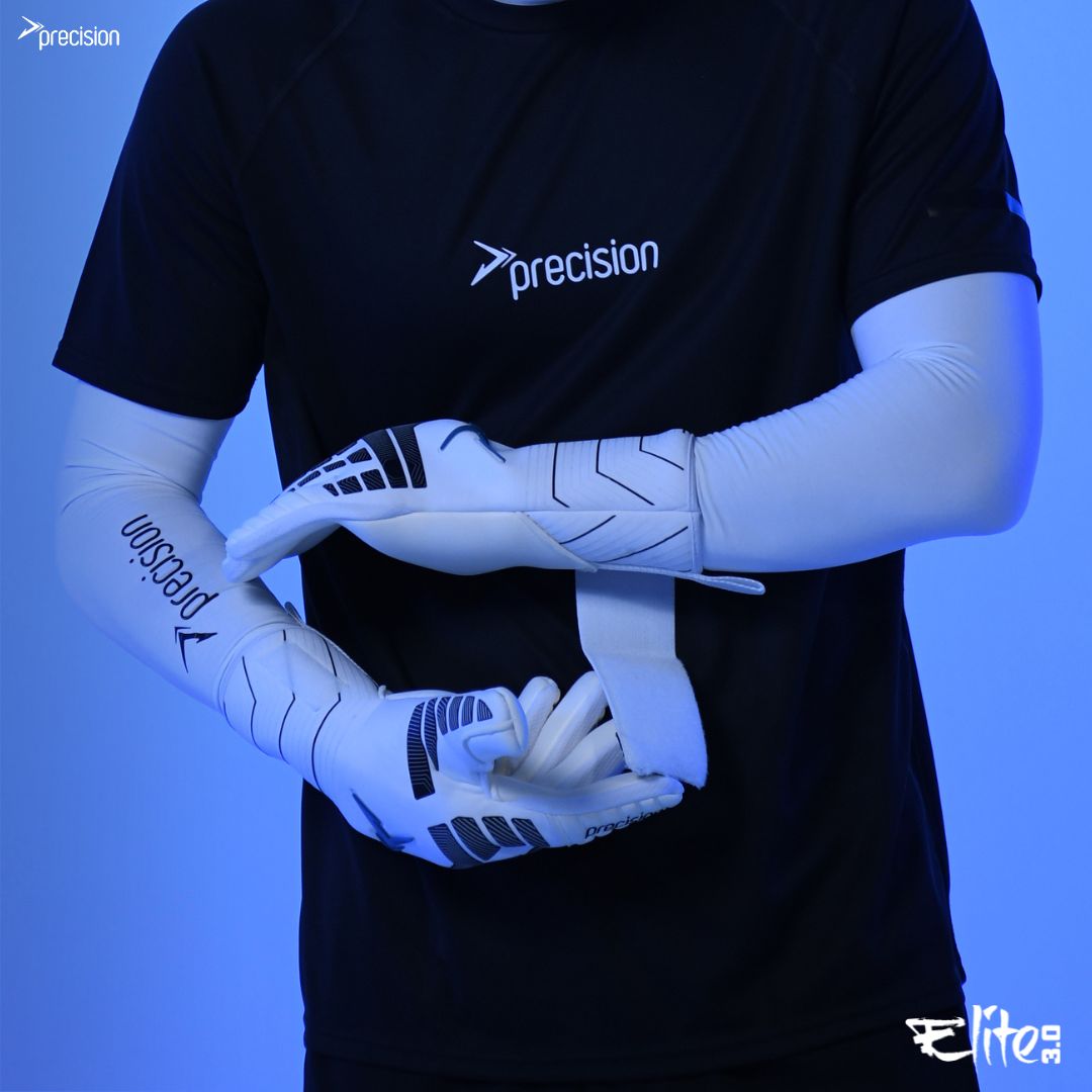 A person in a black Precision shirt stands against a blue background, holding Martin Berrill Sports Precision Elite 3.0 Contact Goalkeeper Gloves in a ready position.