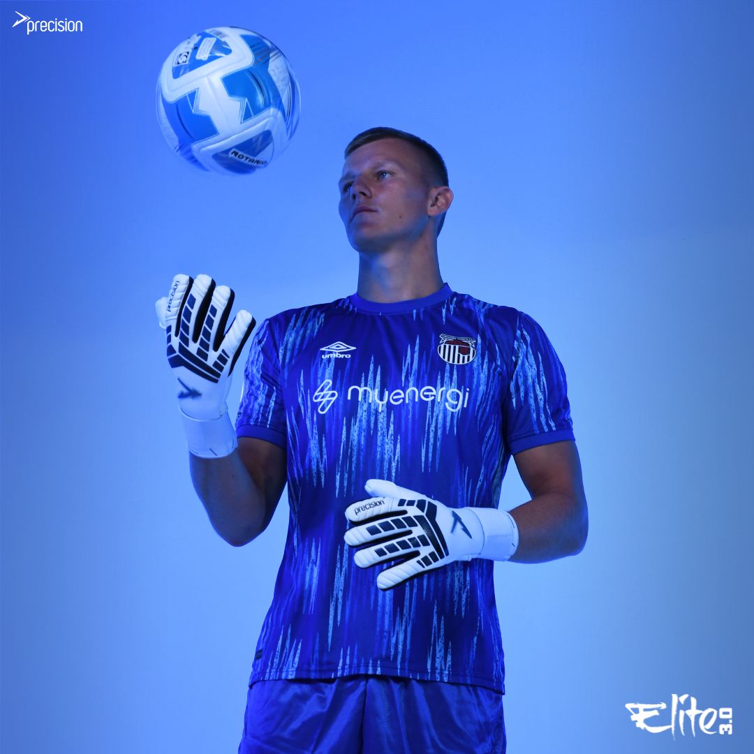 A male football goalkeeper in a blue and purple kit tosses a soccer ball, showcasing his Precision Training Elite 3.0 Giga Goalkeeper Gloves with elite grip. Logos are visible on his shirt and gloves, set against a blue background.
