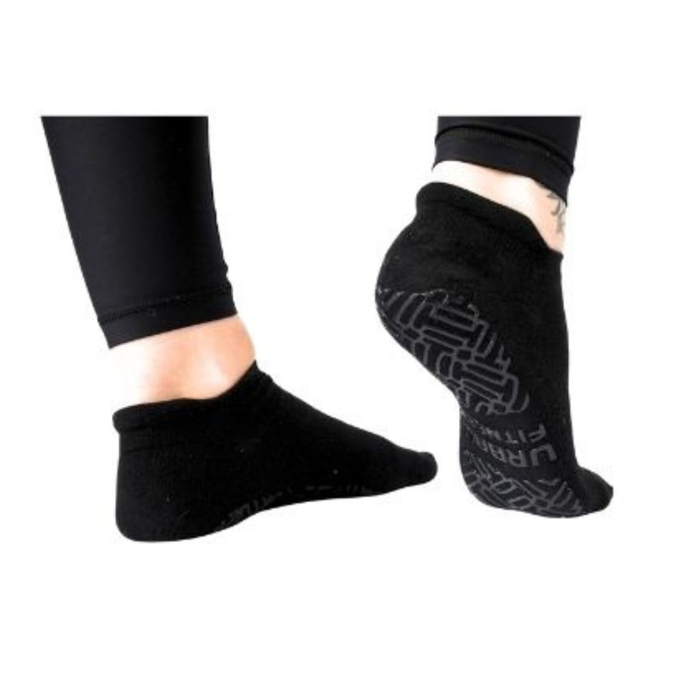 A pair of feet in Urban Fitness Yoga Pilates Fitness Low Rise Grip Socks in black—one flat, one raised to show the non-slip sole—paired with black leggings. Ideal for yoga or lounging with comfort, style, and secure grip.