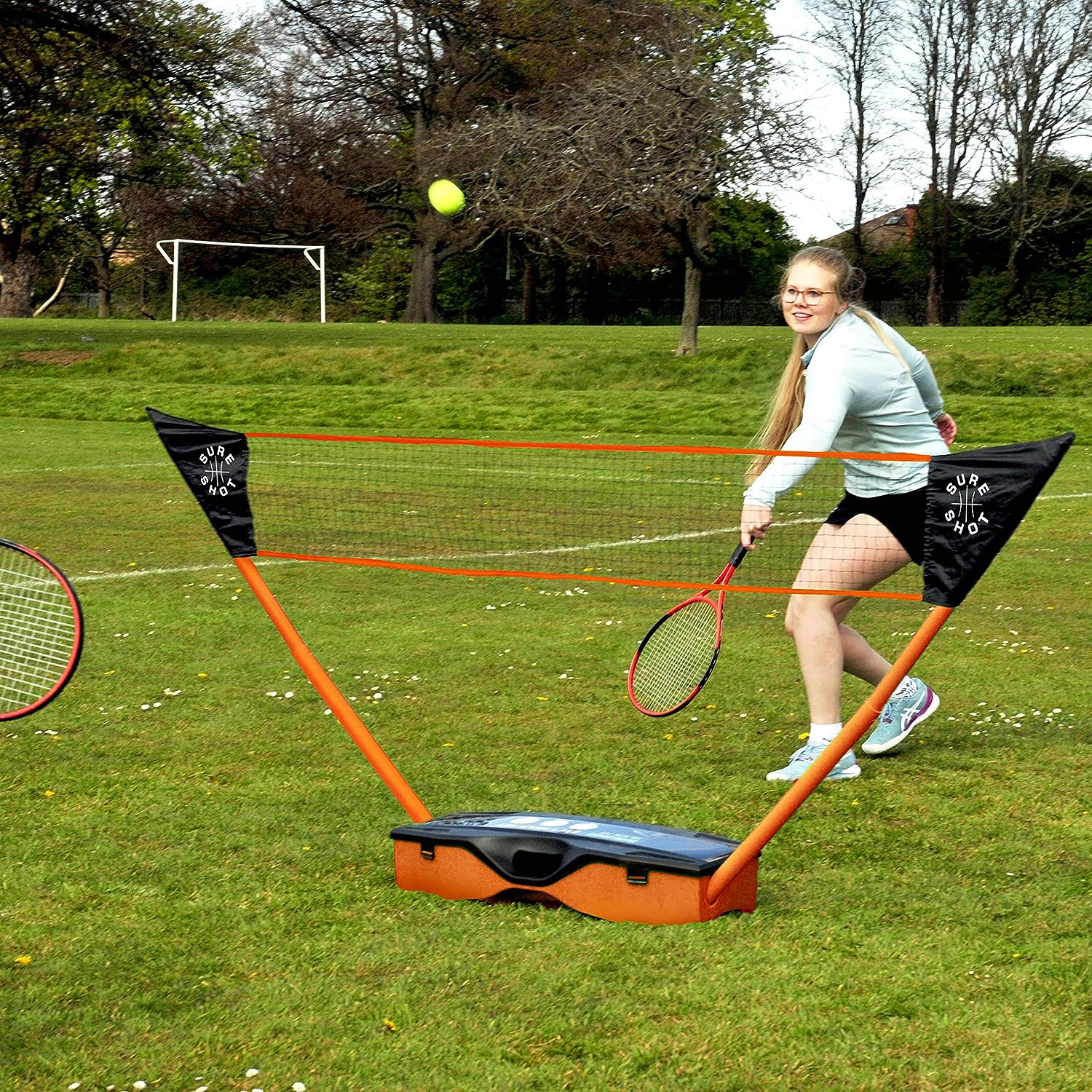 A woman in athletic wear plays badminton on grass, swinging her racket at a shuttlecock with the Sure Shot Quick Fit 3 in 1 Garden Games set by Sure Shot; another racket and trees are visible in the background.