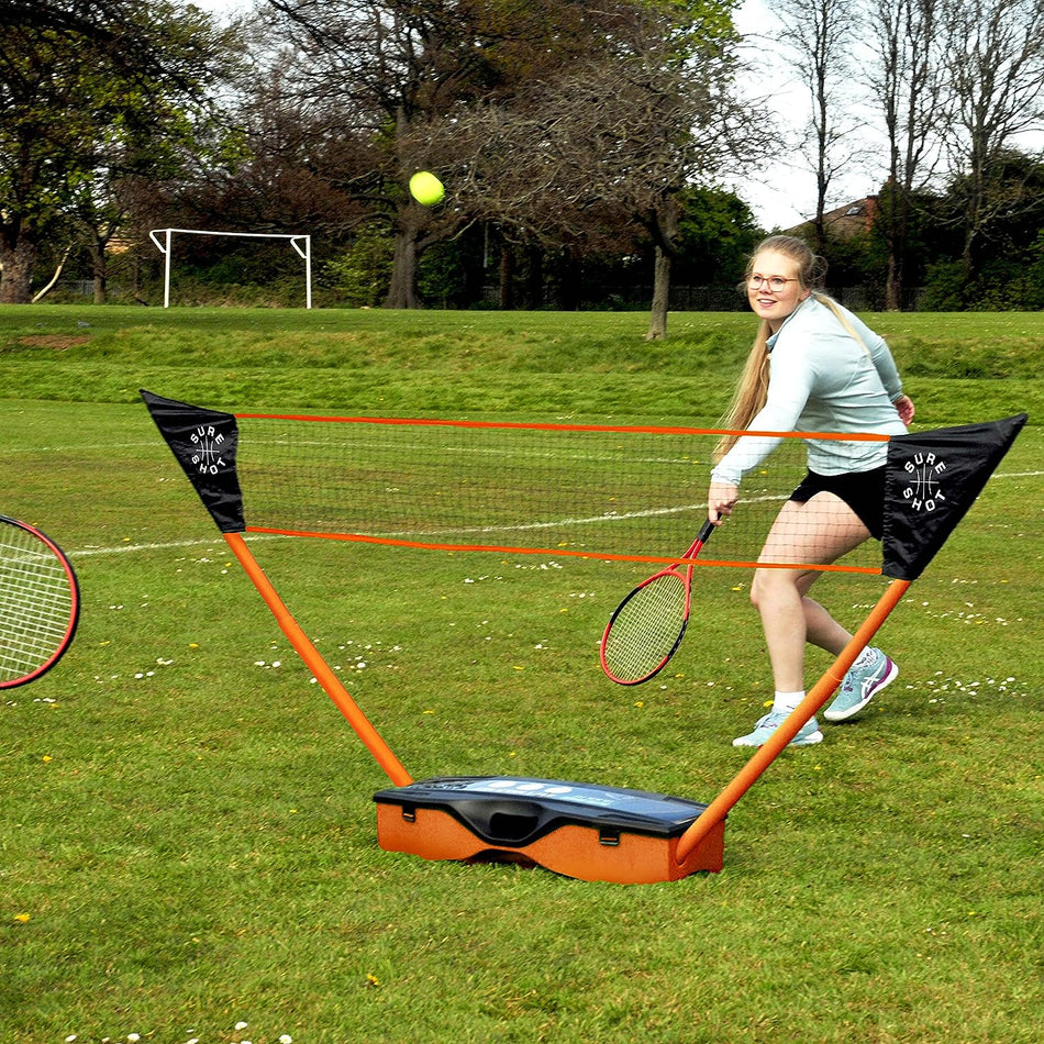 A woman in athletic wear plays badminton on grass, swinging her racket at a shuttlecock with the Sure Shot Quick Fit 3 in 1 Garden Games set by Sure Shot; another racket and trees are visible in the background.