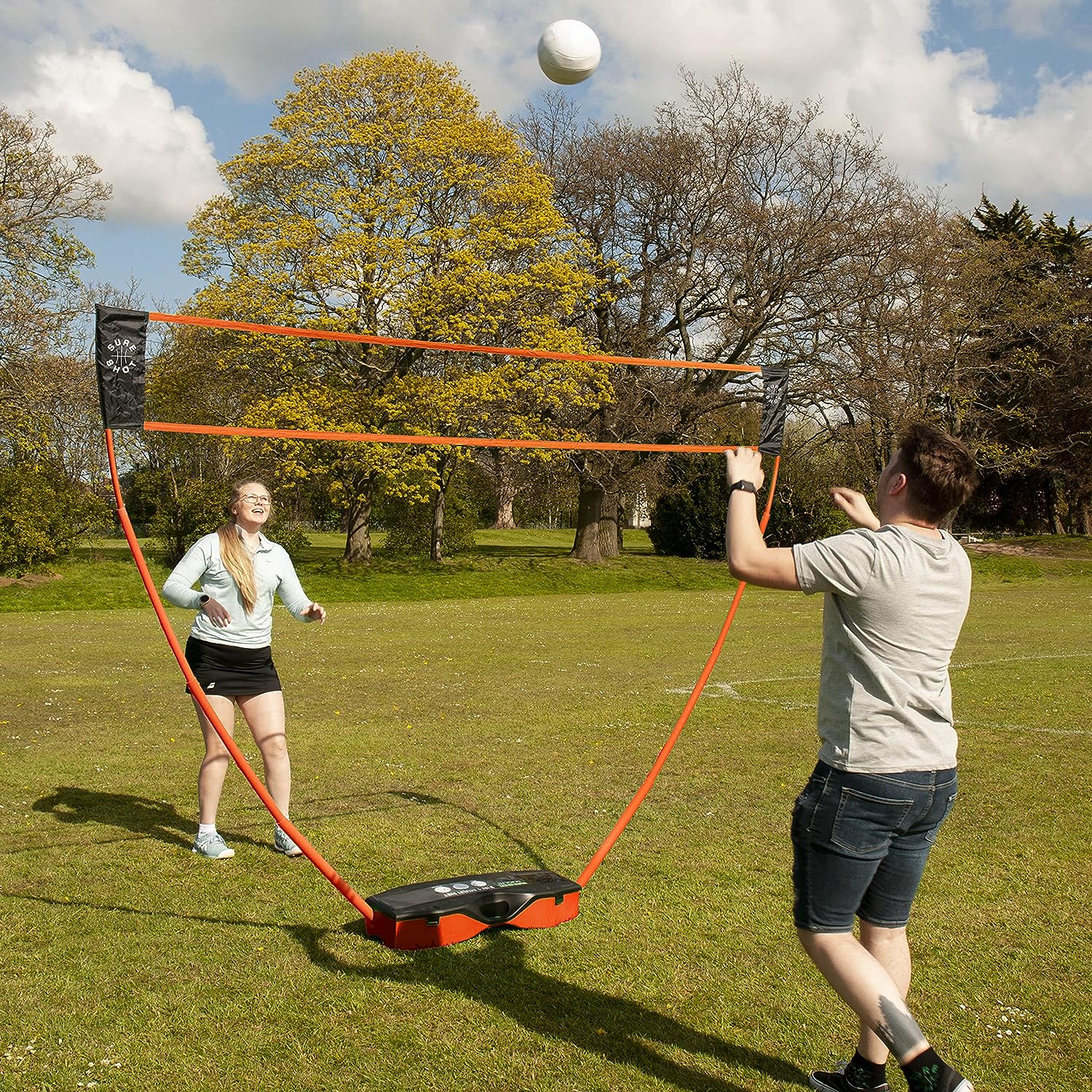 Two people play badminton on grass using the Sure Shot Quick Fit 3 in 1 Garden Games set by Sure Shot, featuring a quick-fit adjustable net. One prepares to hit the shuttlecock while the other gets ready to return it under a cloudy sky and trees.