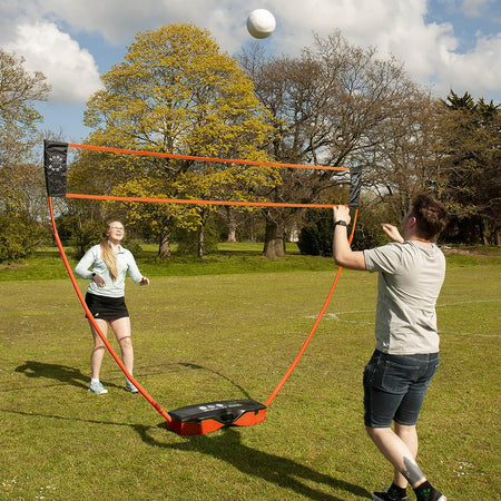 Two people play badminton on grass using the Sure Shot Quick Fit 3 in 1 Garden Games set by Sure Shot, featuring a quick-fit adjustable net. One prepares to hit the shuttlecock while the other gets ready to return it under a cloudy sky and trees.