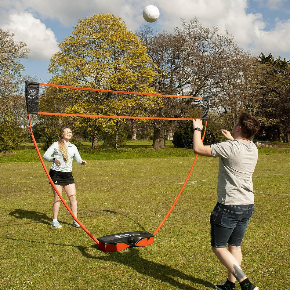 Two people play badminton on grass using the Sure Shot Quick Fit 3 in 1 Garden Games set by Sure Shot, featuring a quick-fit adjustable net. One prepares to hit the shuttlecock while the other gets ready to return it under a cloudy sky and trees.