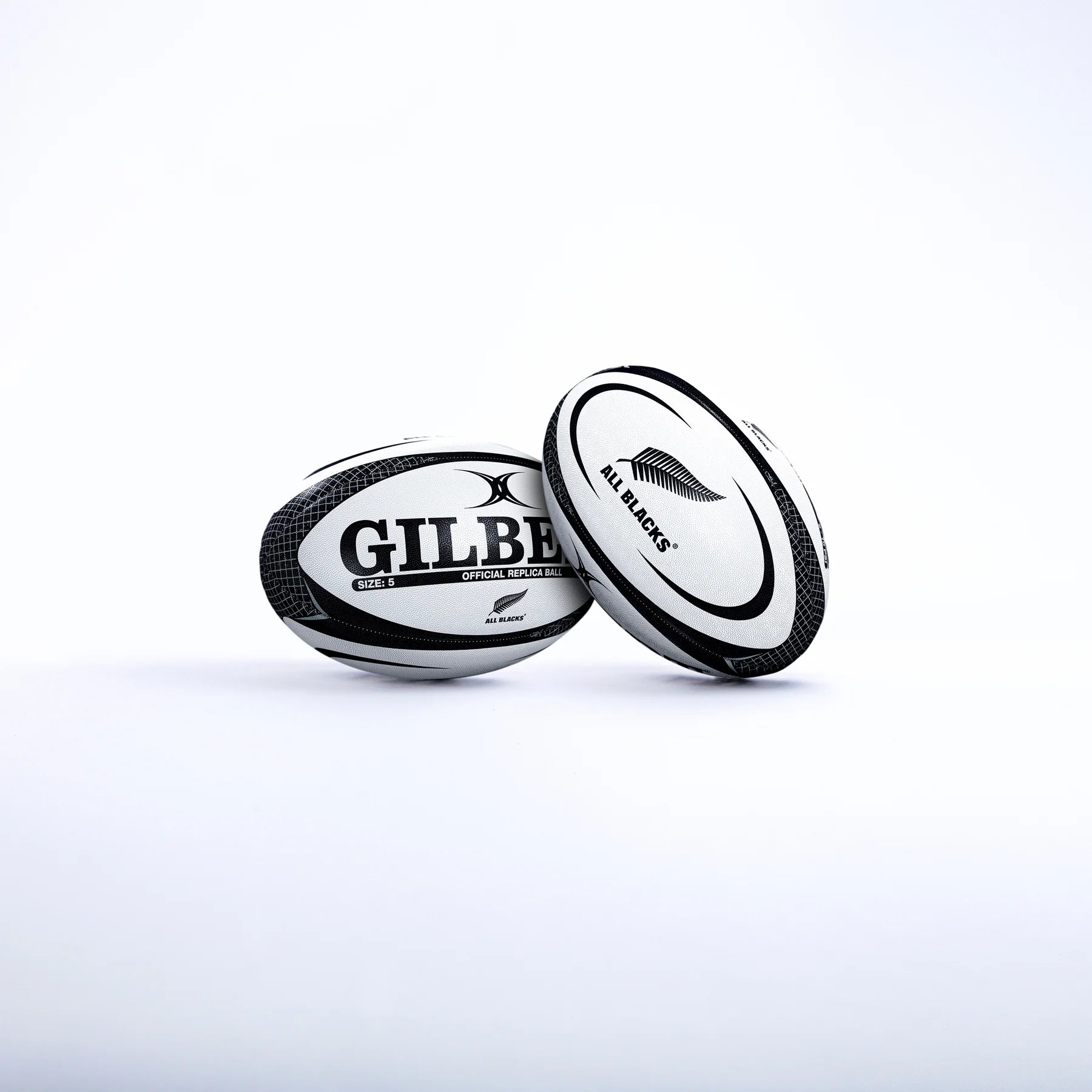 Two Gilbert rugby balls—one with the GILBERT logo and the other a Size 5 All Blacks Replica Rugby Ball featuring the New Zealand All Blacks silver fern—are set against a plain white background.