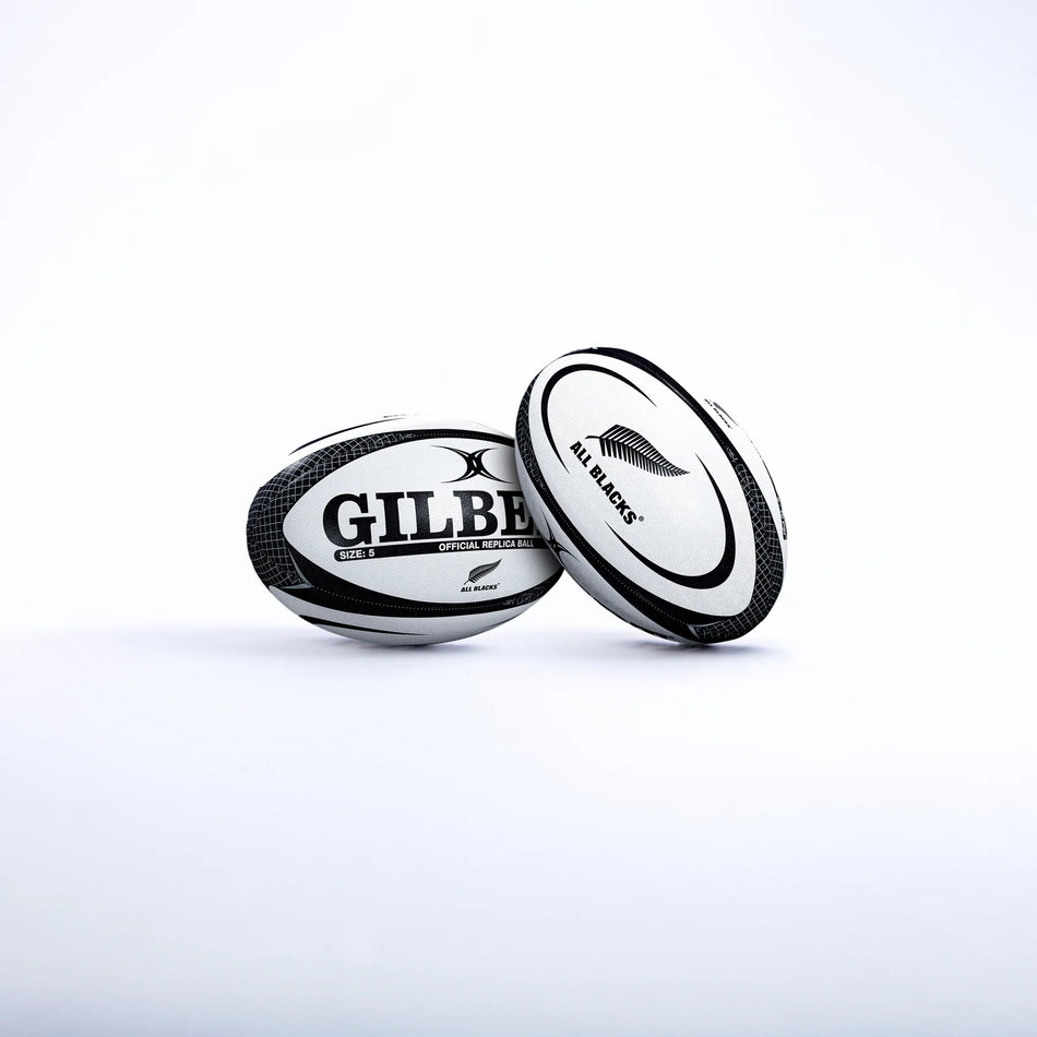 Two Gilbert rugby balls—one with the GILBERT logo and the other a Size 5 All Blacks Replica Rugby Ball featuring the New Zealand All Blacks silver fern—are set against a plain white background.
