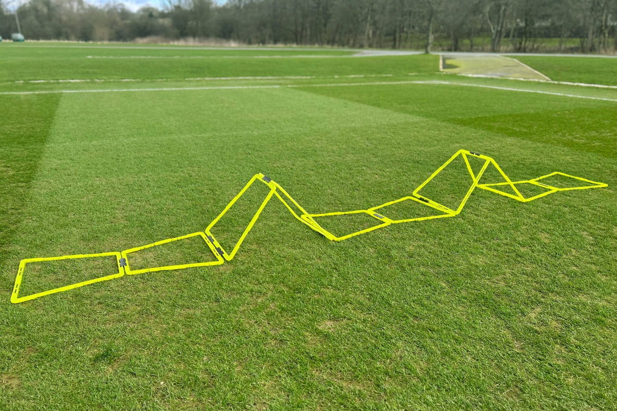 The Samba Pro Agility Trapeze Set by Samba Sports is arranged in a zigzag on a grassy sports field, with trees and a path in the background.