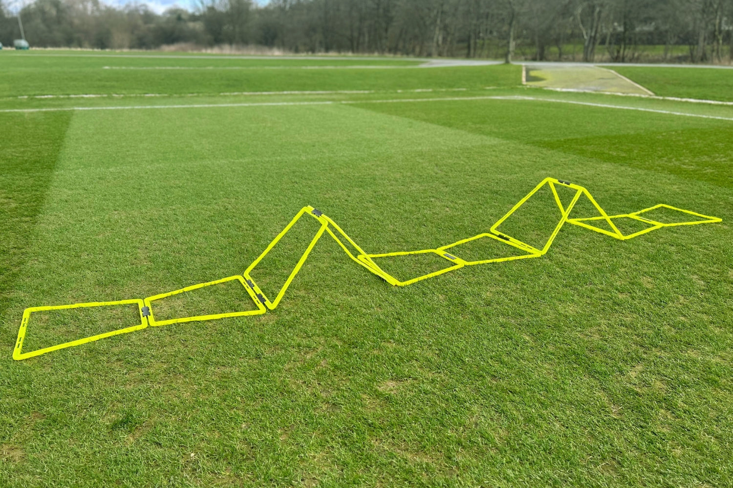 The Samba Pro Agility Trapeze Set by Samba Sports is arranged in a zigzag on a grassy sports field, with trees and a path in the background.