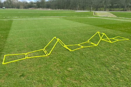 The Samba Pro Agility Trapeze Set by Samba Sports is arranged in a zigzag on a grassy sports field, with trees and a path in the background.