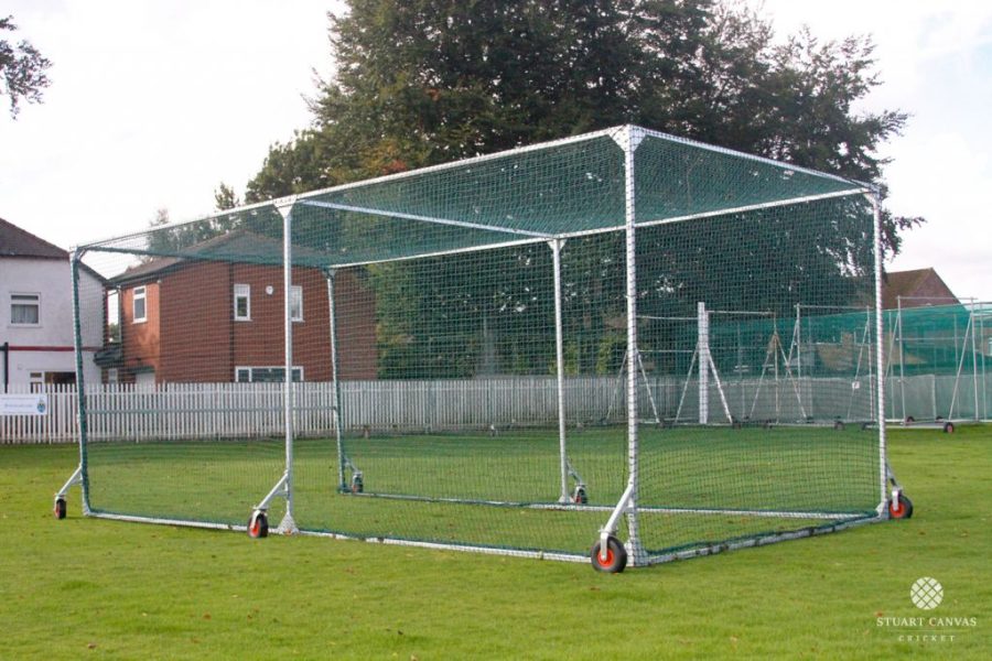 The Stuart Canvas County Batting Cage, a mobile cricket net with a metal frame and wheels, is set up on grass, providing a convenient training enclosure amidst residential buildings and trees.