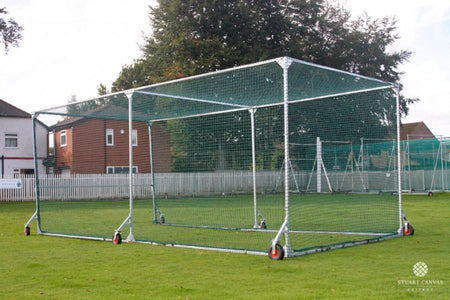 The Stuart Canvas County Batting Cage, a mobile cricket net with a metal frame and wheels, is set up on grass, providing a convenient training enclosure amidst residential buildings and trees.