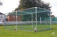 The Stuart Canvas County Batting Cage, a mobile cricket net with a metal frame and wheels, is set up on grass, providing a convenient training enclosure amidst residential buildings and trees.