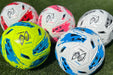 Five Samba Infiniti 4 Max Training Balls by Martin Berrill Sports—two white/blue, two white/pink, and one yellow/blue—rest on green grass. Each has precision-moulded grooves for better control.